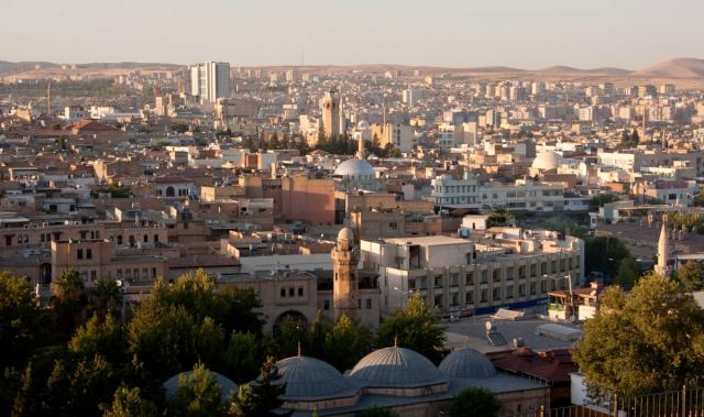 Golden-hour skyline of Şanlıurfa's old town, with domed mosque roofs and a minaret in the foreground against the wider city and surrounding hills.