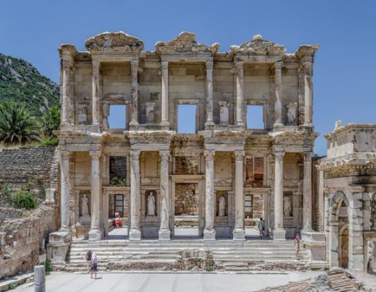The two-storey columned facade of the Library of Celsus at Ephesus under a clear blue sky, with a few visitors on the steps.