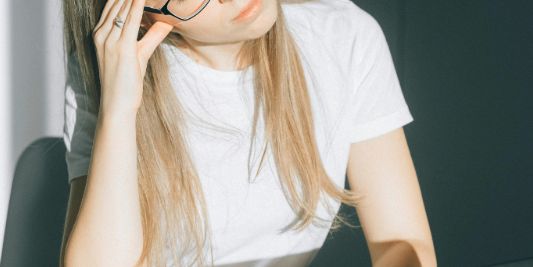 A woman wearing glasses sits at a desk in front of a laptop. She appears tired.