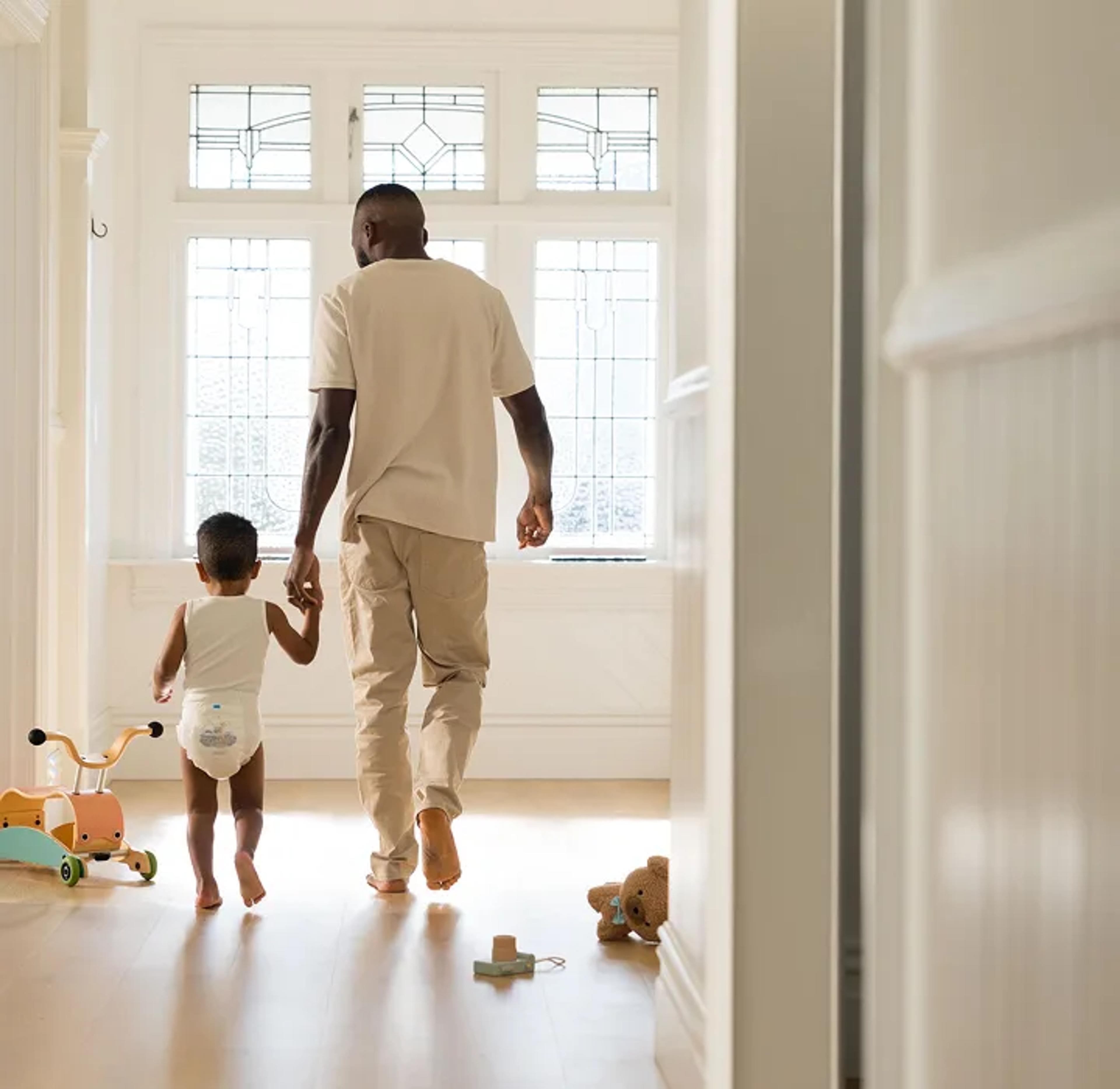 Parent walking hand in hand down a sunlit hallway with a toddler in a Millie Moon diaper