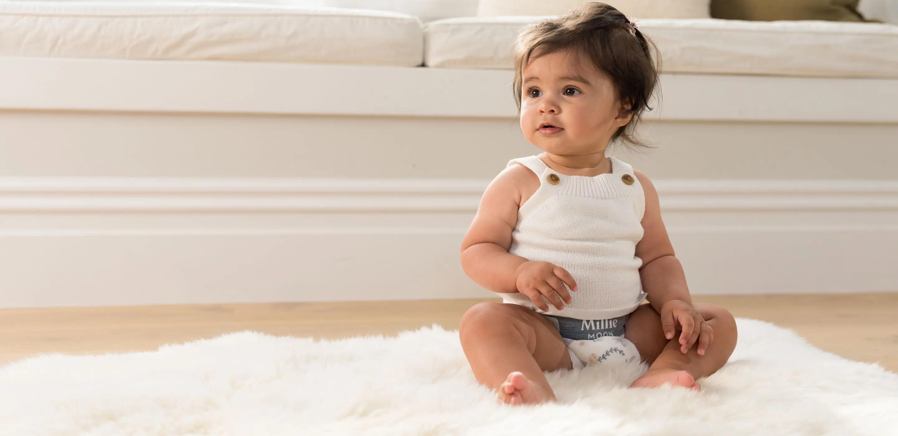 Baby in a knitted vest and a Millie Moon Luxury Diaper sitting on a sheepskin rug