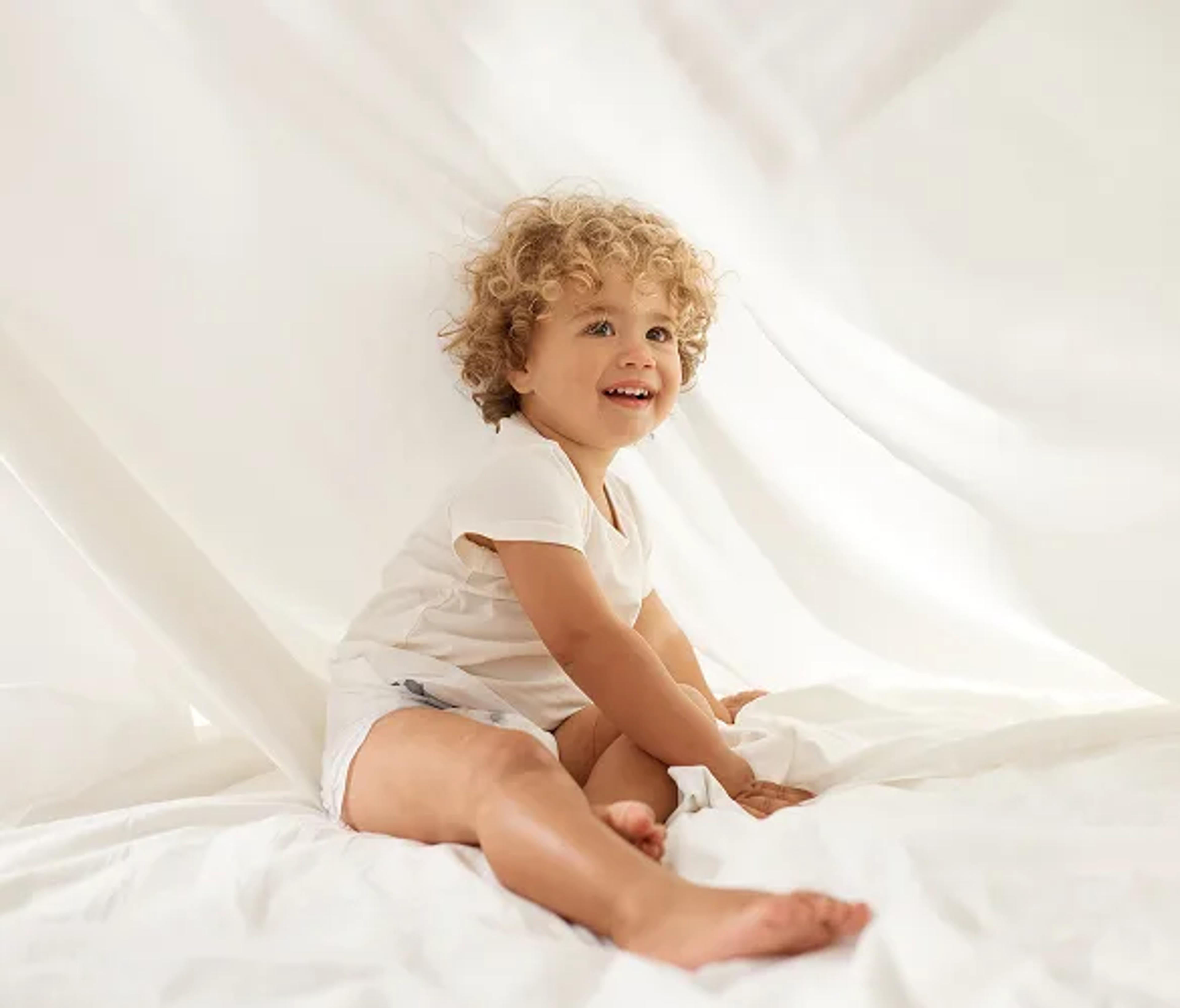 Smiling toddler in a Millie Moon diaper sitting among soft white sheets