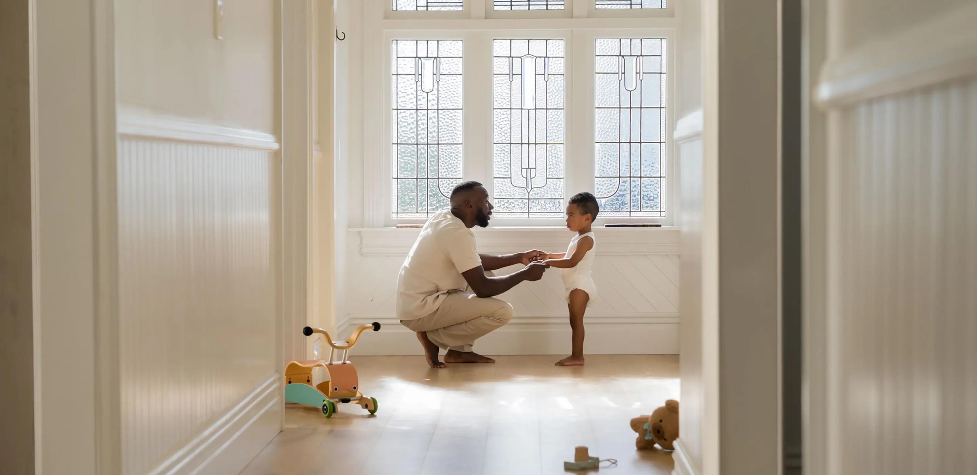 Parent crouching in a sunlit hallway holding the hands of a toddler in a Millie Moon diaper