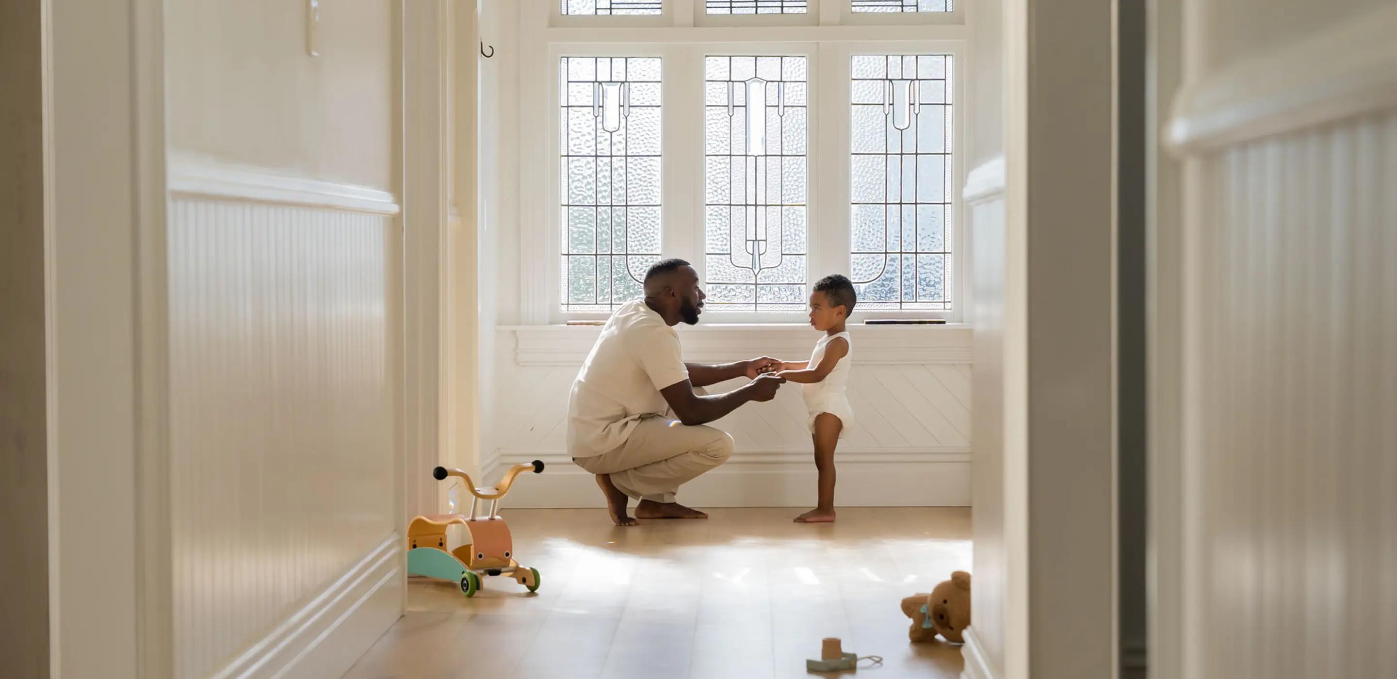 Parent crouching in a sunlit hallway holding the hands of a toddler in a Millie Moon diaper