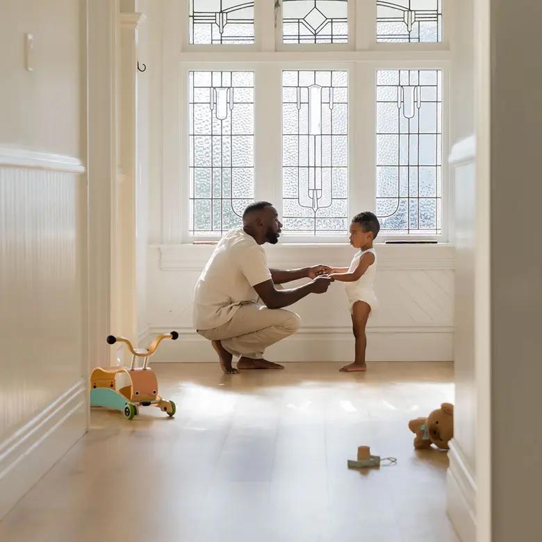 Parent crouching in a sunlit hallway holding the hands of a toddler in a Millie Moon diaper
