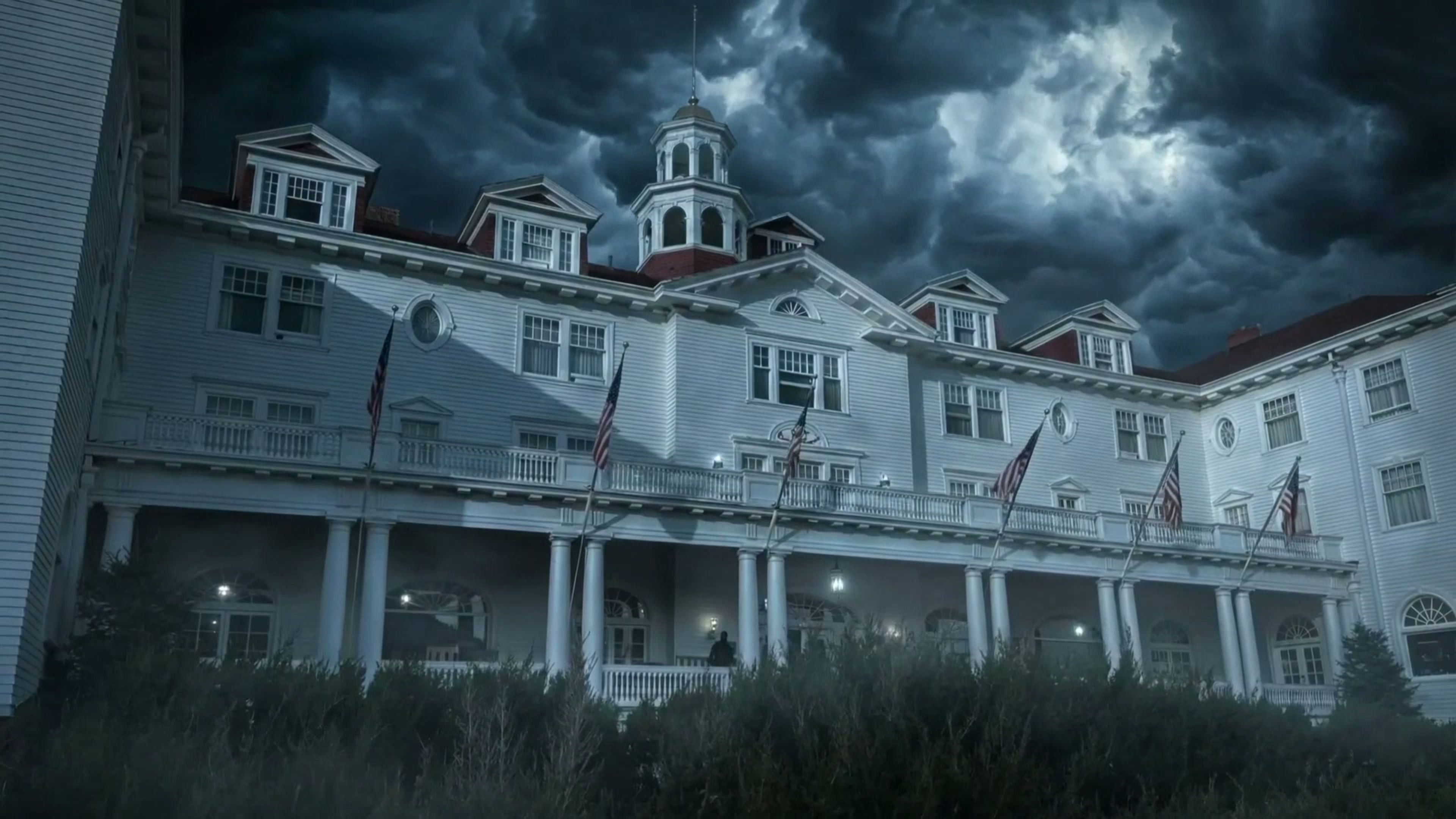 The white wooden Stanley Hotel in Colorado at night under heavy storm clouds, with flags along its long porch and dark bushes in front