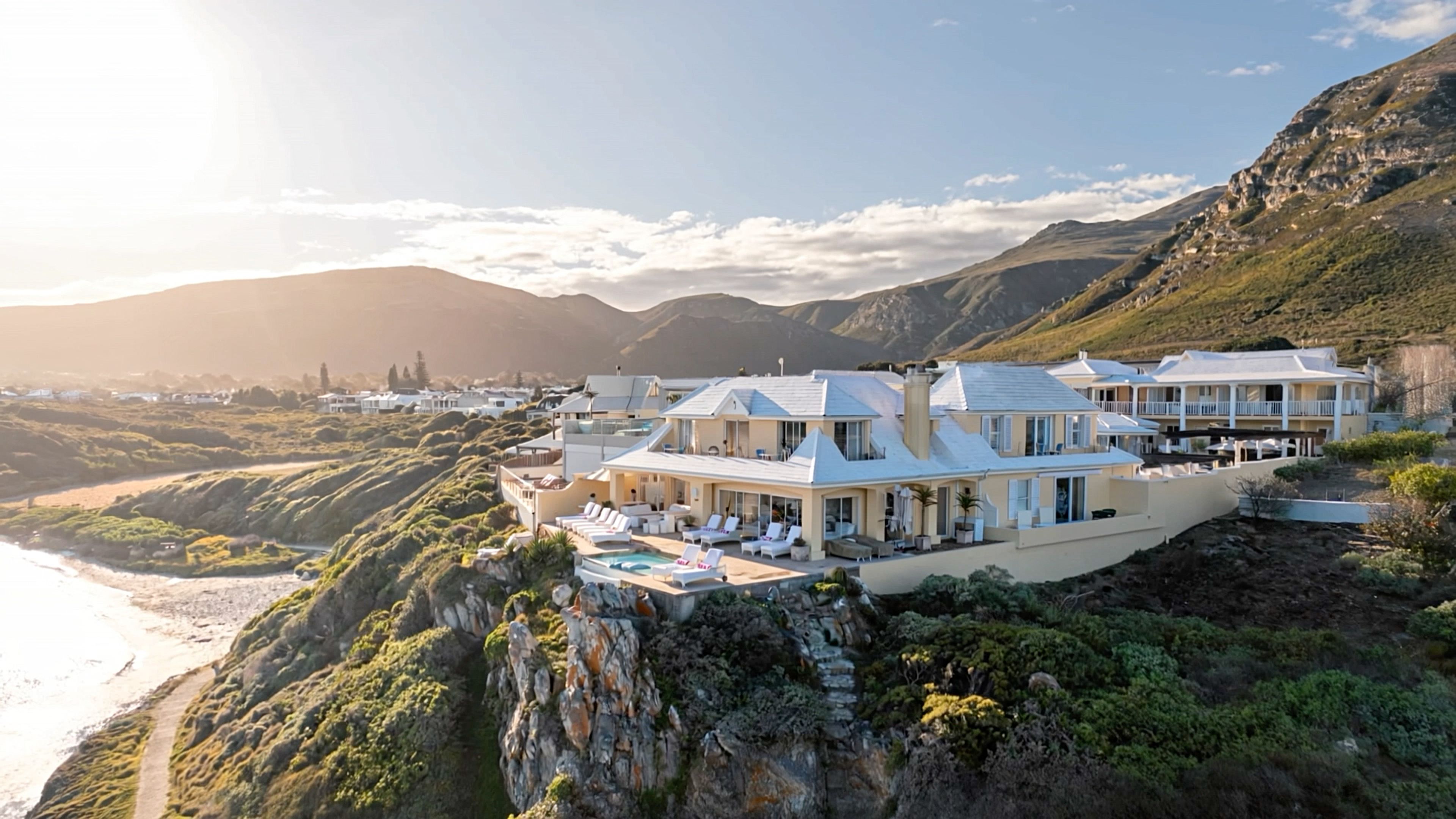 Aerial view of a whitewashed clifftop hotel above a curving sandy beach in Hermanus, South Africa, at golden hour
