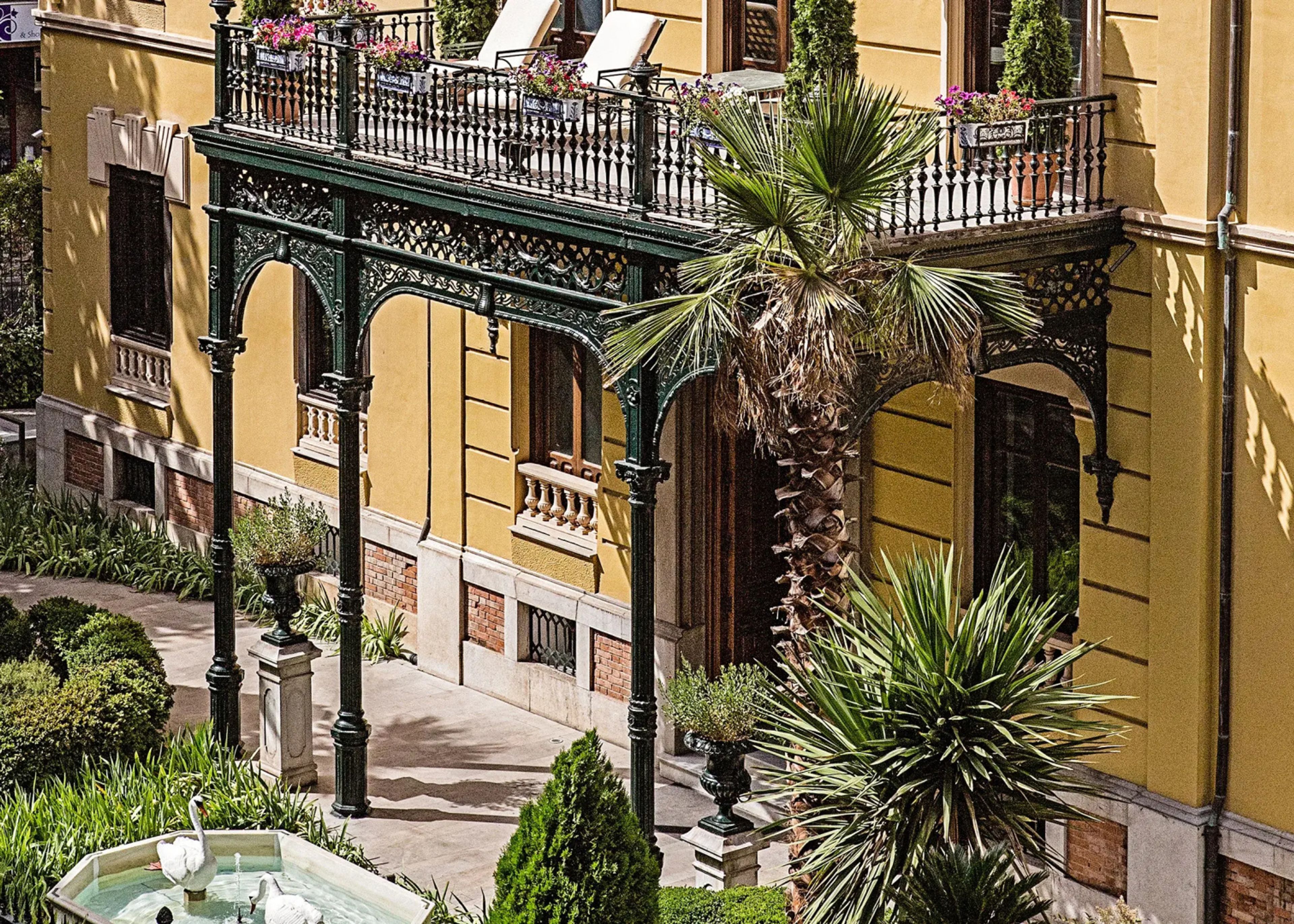Ornate green cast-iron veranda on the yellow facade of a palace hotel in Granada, Spain, with palms and a fountain in the garden below
