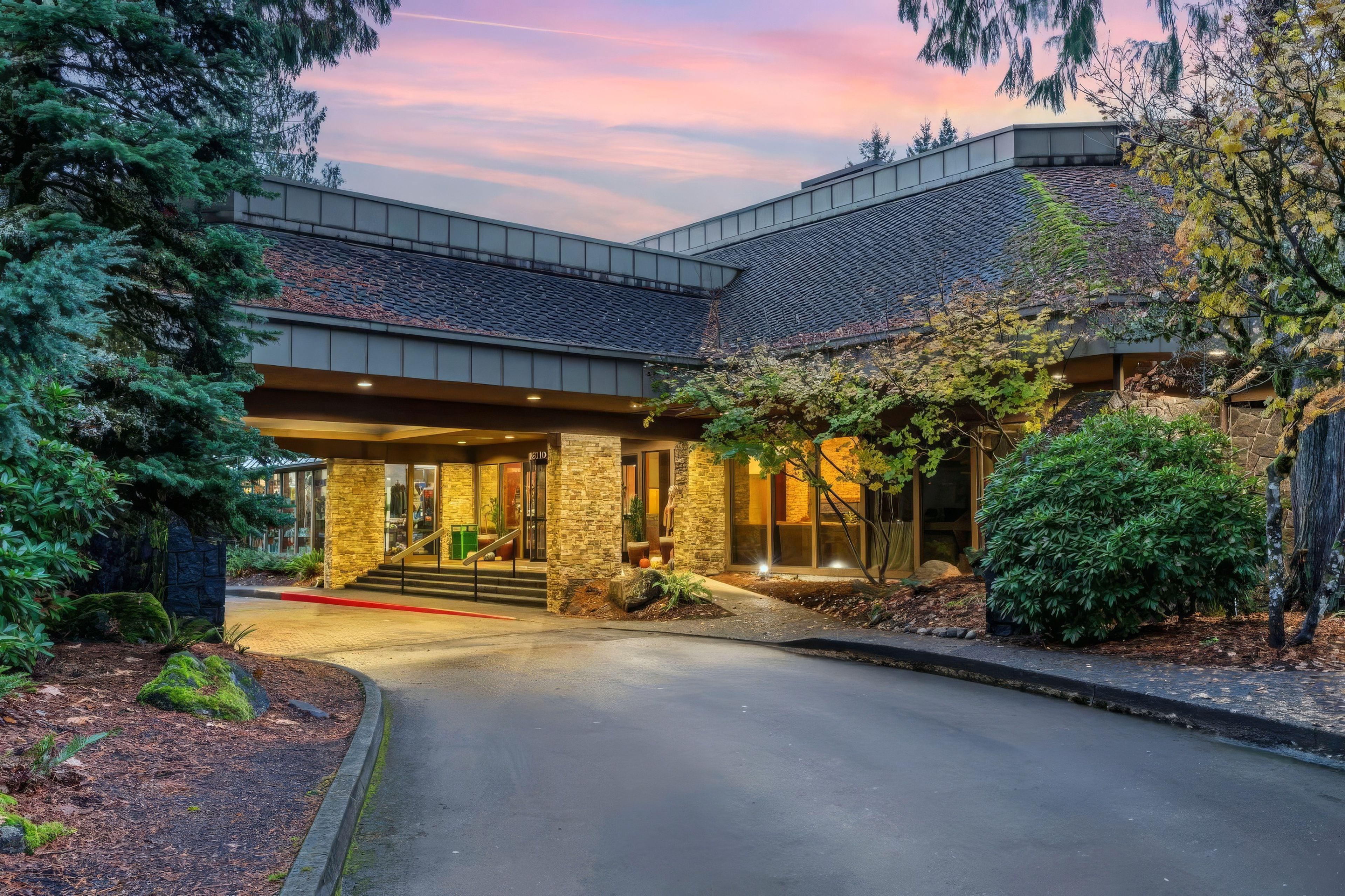 Lit stone and timber porte-cochere entrance of a lodge hotel near Mount Hood, Oregon, framed by conifers under a pink sunset sky