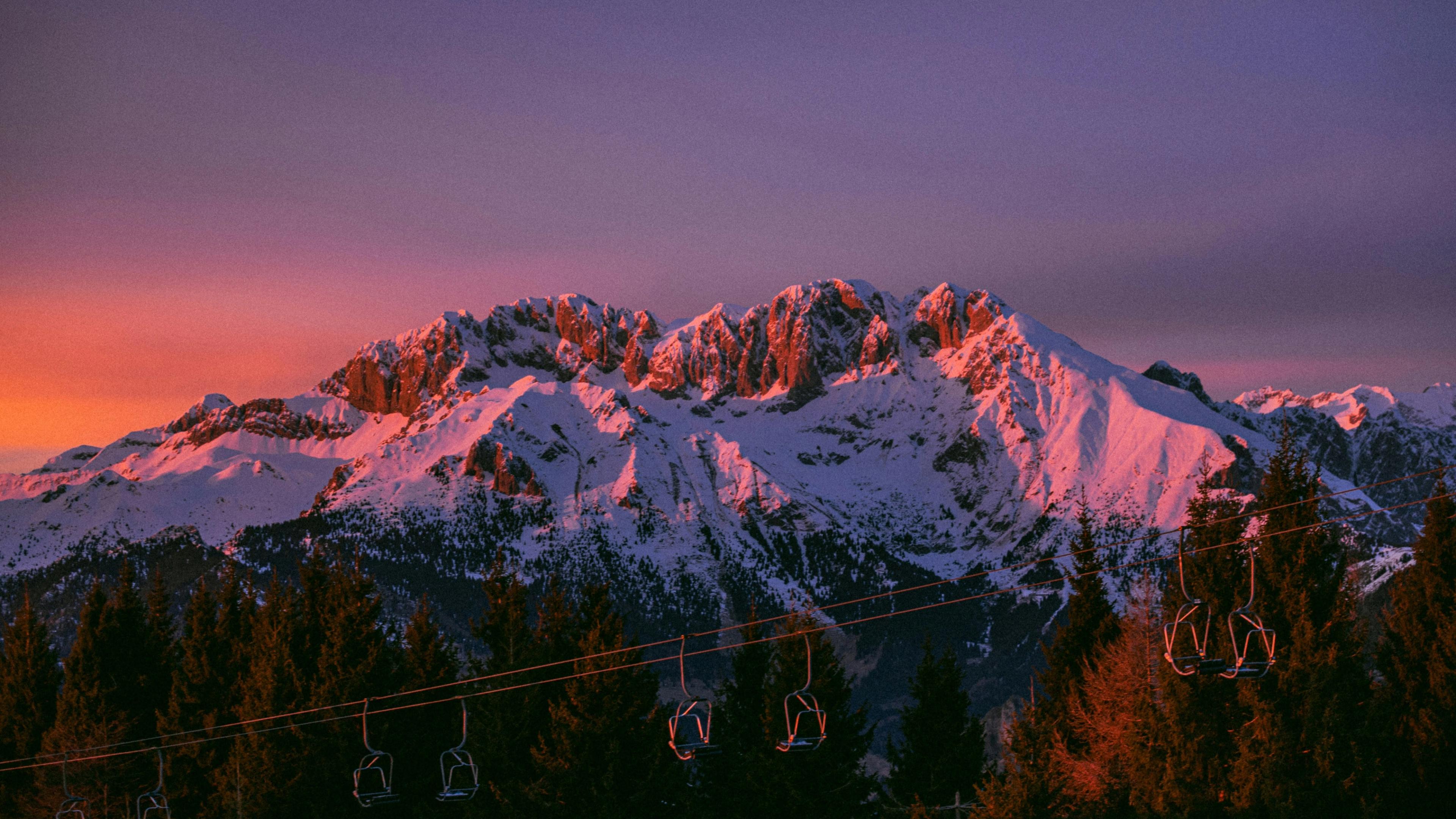 Alpenglow on a snow-covered Alpine ridge at first light, with an empty chairlift crossing the dark conifers below