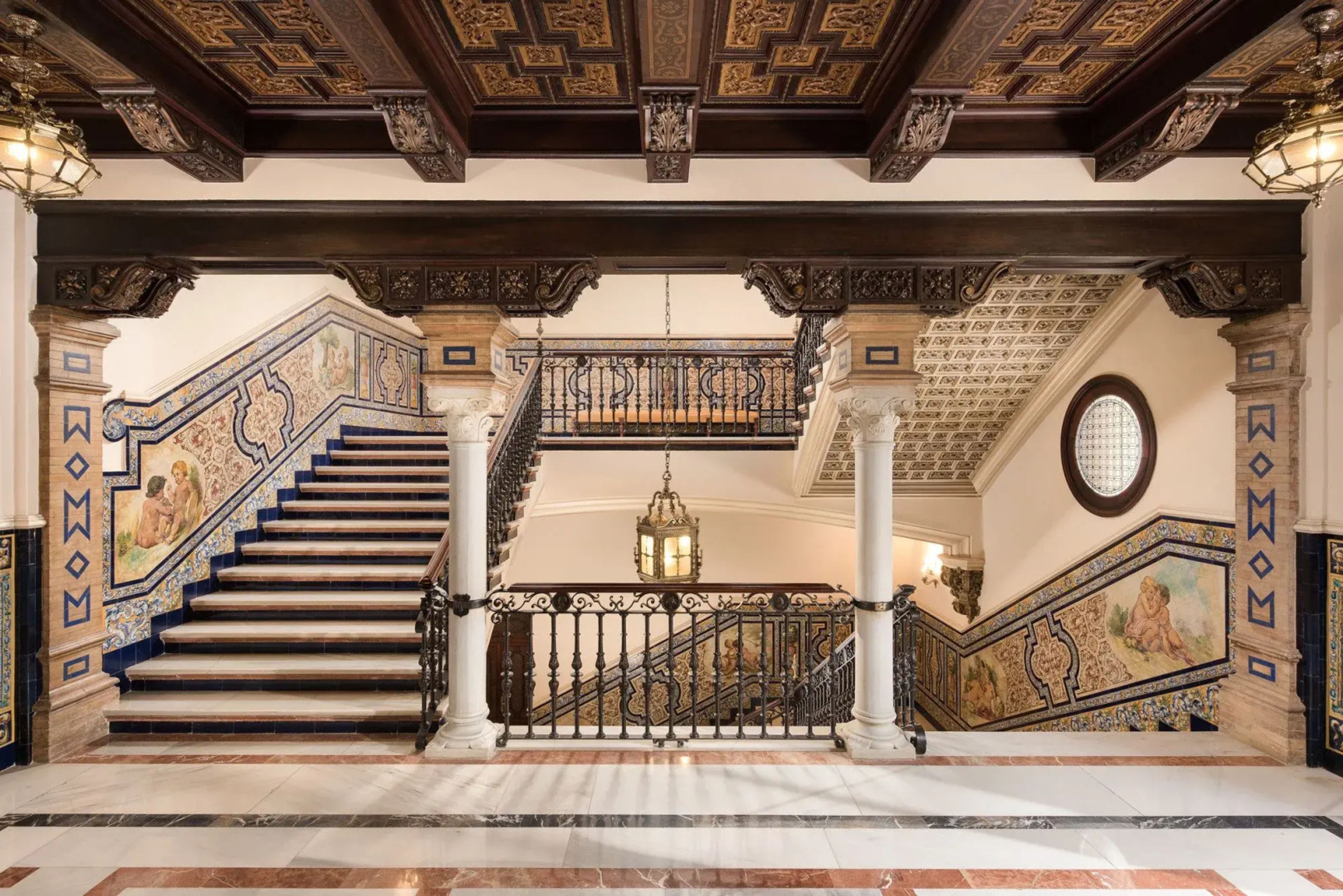 The grand tiled staircase of Hotel Alfonso XIII in Seville, with painted azulejo panels, marble columns and a carved coffered wooden ceiling