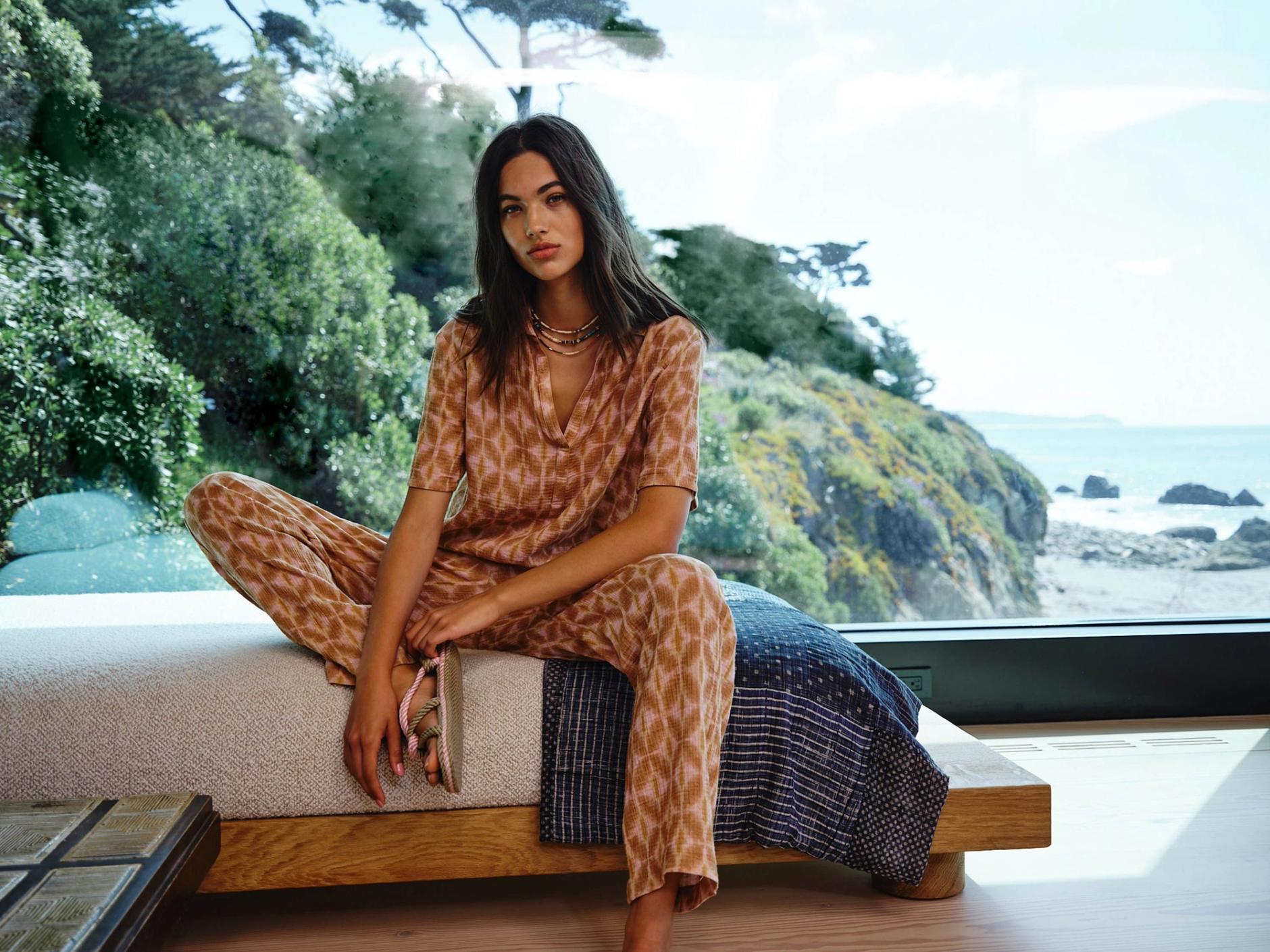 Woman in Xirena outfit sits on a bed by the ocean