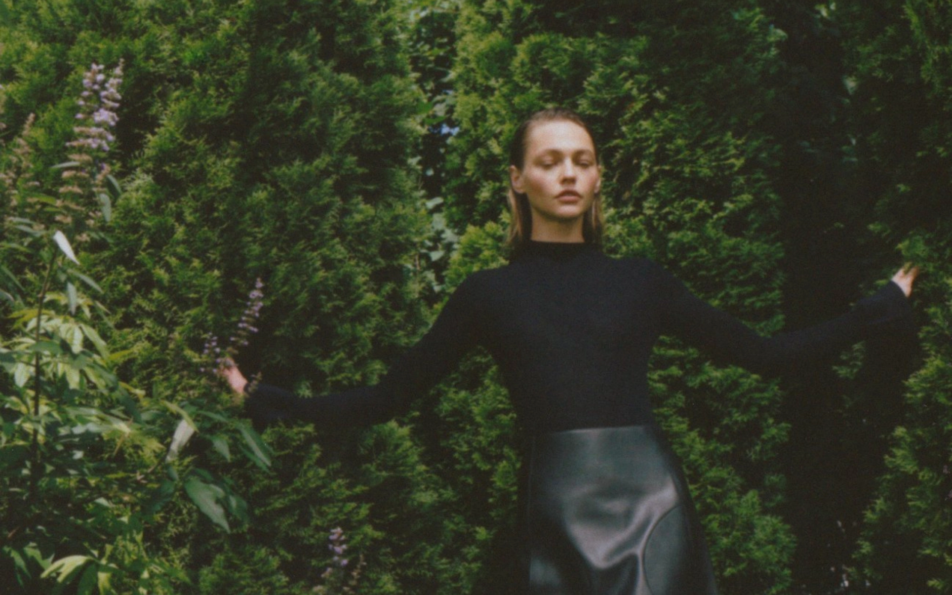 Woman in Khaite turtleneck and leather skirt in front of a large hedge