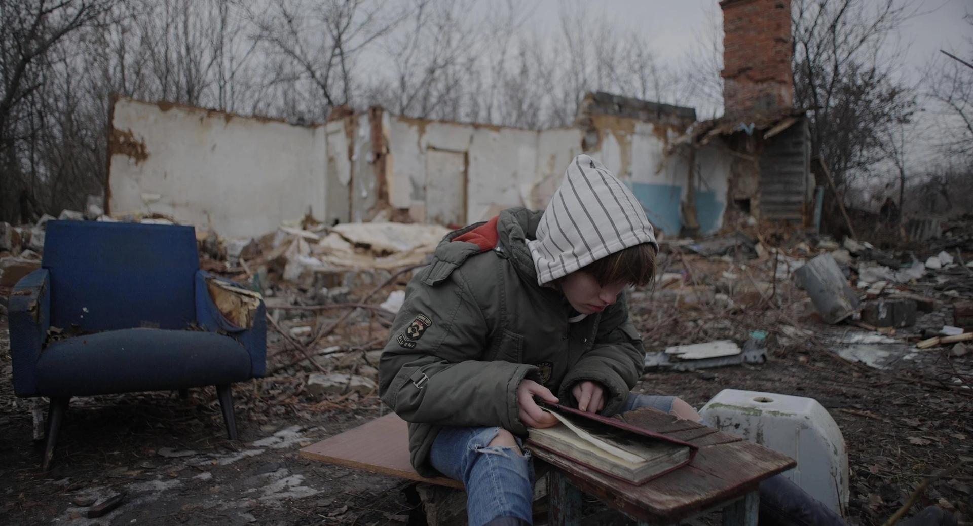A still from the film Boney Piles (2022) by Taras Tomenko, showing a boy in eastern Ukraine, sitting and reading a book in what was once his home Courtesy of Taras Tomenko