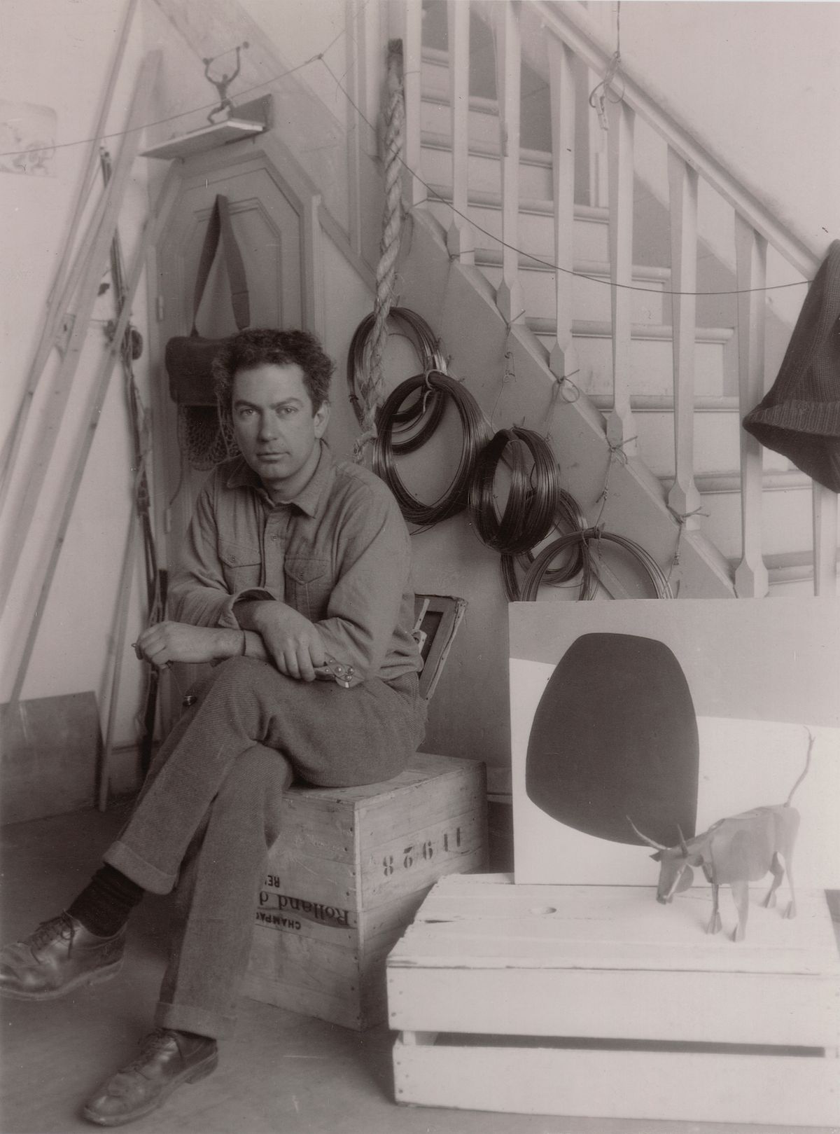Calder with Old Bull and an oil painting, 7 Villa Brune studio, Paris, November 1930. Coils of wires that he used in his mobile sculptures hand behind him Photo: Thérèse Bonney © Estate of Thérèse Bonney / Bancroft Library, University of California at Berkeley © 2021 Calder Foundation, New York / Artists Rights Society (ARS), New York