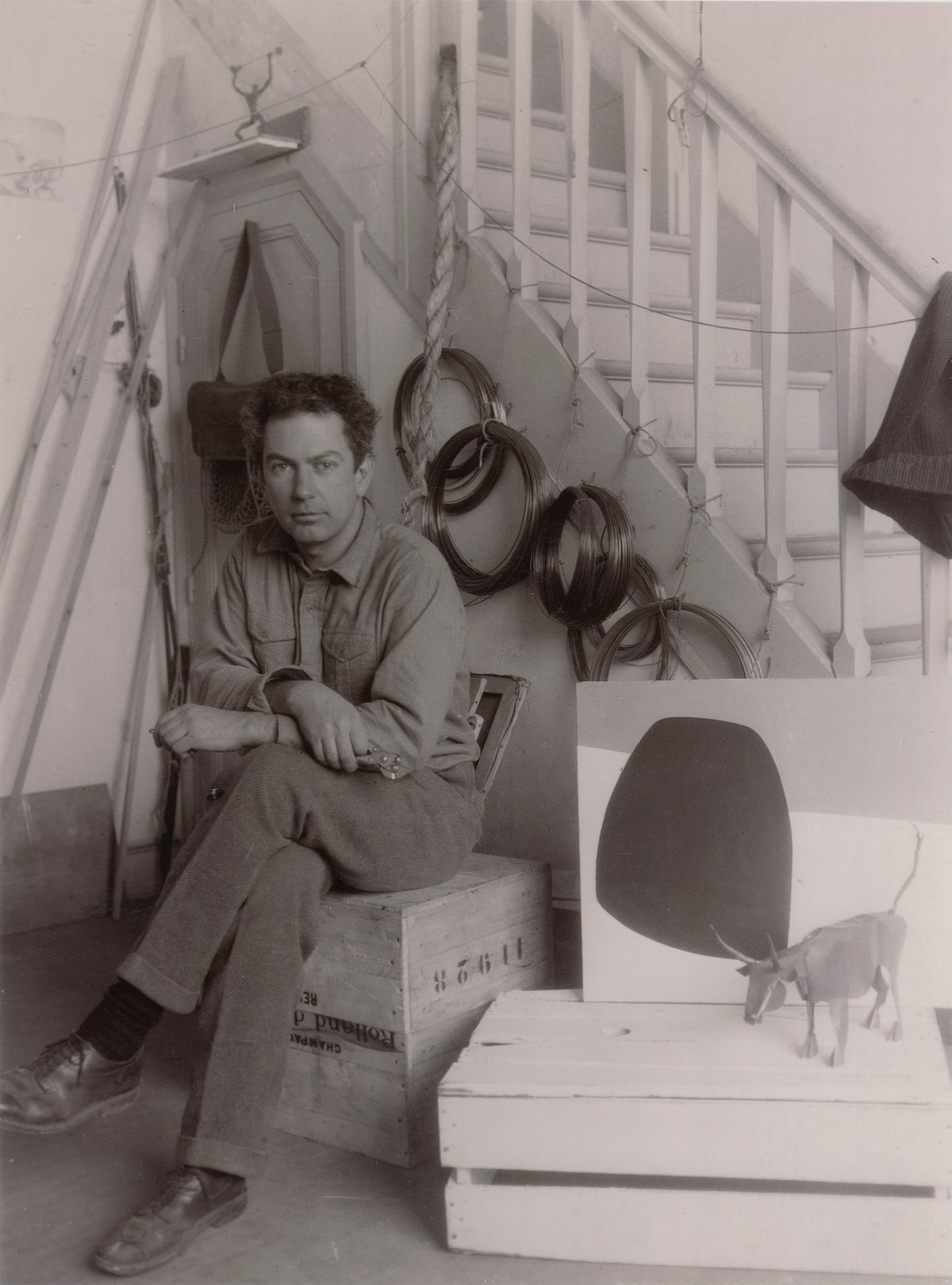 Calder with Old Bull and an oil painting, 7 Villa Brune studio, Paris, November 1930. Coils of wires that he used in his mobile sculptures hand behind him Photo: Thérèse Bonney © Estate of Thérèse Bonney / Bancroft Library, University of California at Berkeley © 2021 Calder Foundation, New York / Artists Rights Society (ARS), New York