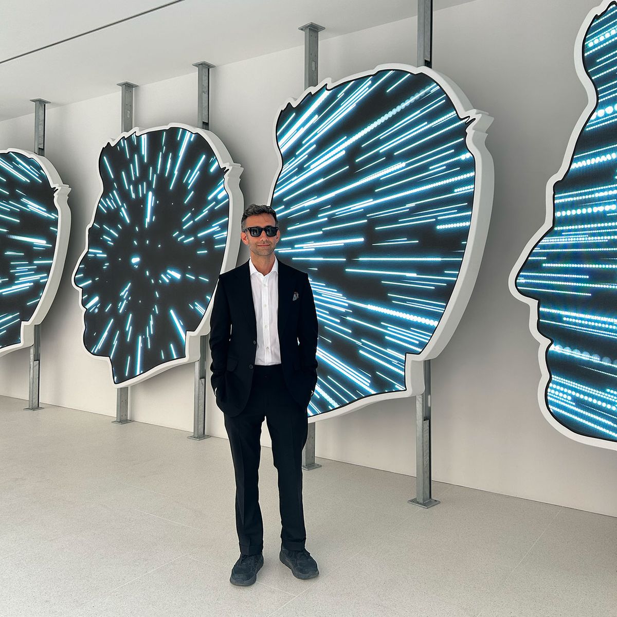 Alex Israel at Gagosian, Davies Street, in front of some of the seven screens—based on his profile-in-shades logo—on which REMEMBR projects videos generated using AI from user's smartphone camera rolls Photograph: The Art Newspaper