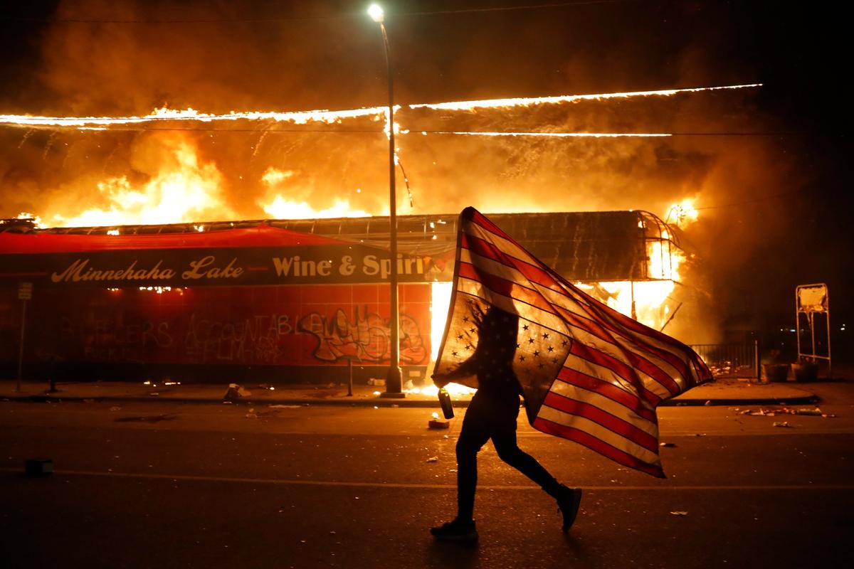 A protester carries a US flag upside down, a sign of distress, next to a burning building Thursday, 28 May 2020, in Minneapolis. Protests over the death of George Floyd, a black man who died in police custody Monday, broke out in Minneapolis for a third straight night AP Photo/Julio Cortez