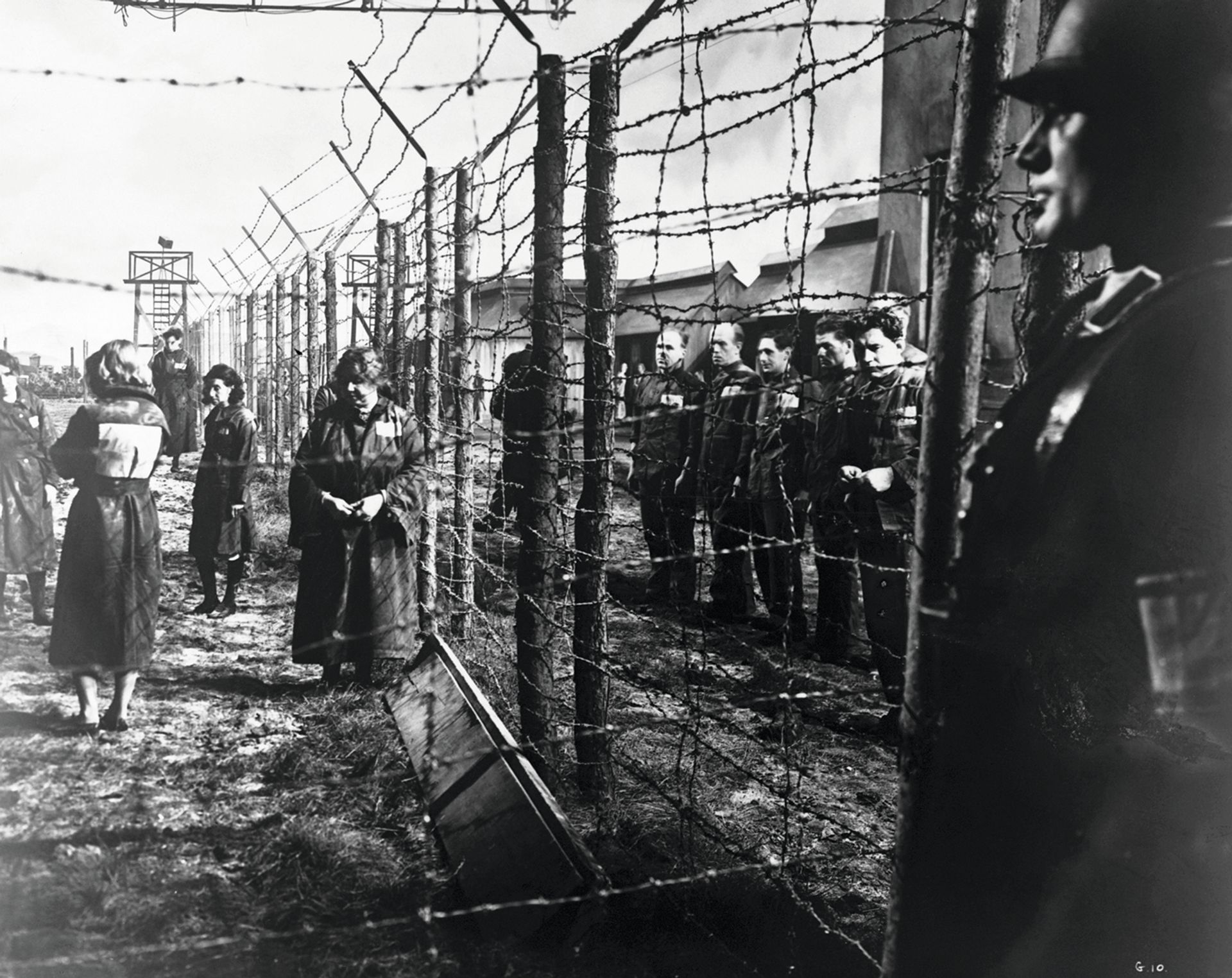 An electrified barbed-wire fence separates male and female prisoners at a German concentration camp. A Nazi guard keeps watch in the foreground. The inmates appear to be in relatively good health at this point in their internment, indicating they may have arrived recently at the camp © Hulton-Deutsch Collection/CORBIS/Corbis via Getty Images)