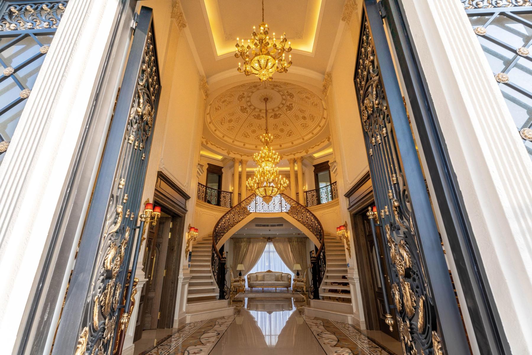 A double-height entrance hall with twin curving staircases, crystal chandeliers, and a gilded coffered dome above a polished marble floor.