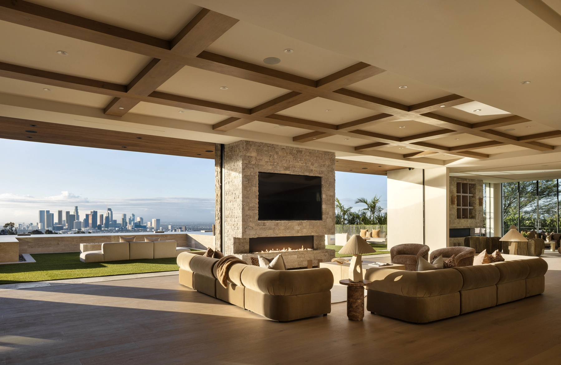 A living room under a coffered timber ceiling, opening through a full glass wall to a terrace and a distant city skyline.
