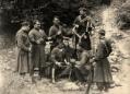 Group of men wearing traditional Georgian dress and holding rifles, gathered outdoors on rocky terrain.
