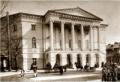 Historic Tiflis Theological Seminary façade with columned portico and arched arcade; horse carriages and pedestrians on the street.