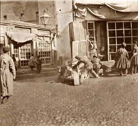 Photograph showing several people working and gathering near a shopfront or workshop in the Tiflis bazaar, with wooden boards and tools visible.