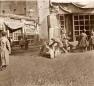 Photograph showing several people working and gathering near a shopfront or workshop in the Tiflis bazaar, with wooden boards and tools visible.