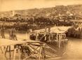 Large crowd gathered around a damaged wooden bridge structure over a river in Tiflis, with city buildings visible in the background.