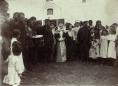Bride in traditional attire standing among men, women, and children outside a church during a wedding ceremony, with a drummer visible.