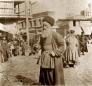 An elderly man standing in a cobbled bazaar street in Tiflis, with market activity and shops visible in the background.
