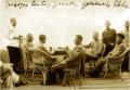 Historic photograph showing a group of men seated and standing around a table on a wooden terrace, with wicker chairs and handwritten text visible above.