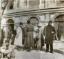 Group of men standing and sitting in front of a large building with arched windows in Tiflis.