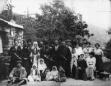 Group of men, women, and children posed outdoors in front of a stone building