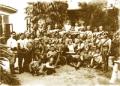 A large group of armed men posing outdoors in front of a building, some standing and others seated, holding rifles and sabers.