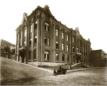 Large multi-storey brick institutional building on a sloping cobblestone street with a few people seated in front.