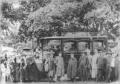 Large crowd surrounding an early automobile with seated passengers beneath trees outdoors.