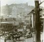 Market square in Tiflis with horse-drawn carts, pedestrians, and buildings, with Narikala fortress visible on the hill.