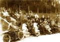 Gelatin silver photograph showing a large seated and standing group of men and women outdoors in a wooded park setting, including a motorcycle in the foreground, Borjomi, 1920.