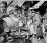 A fish seller cleaning fish at a market stall while several men stand around watching in a bazaar street.