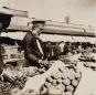 Photograph showing a fruit vendor standing behind baskets of fruit at a market stall in the Tiflis bazaar, late 19th century.