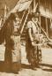 Sepia-toned photograph showing two women in traditional regional dress standing outdoors near a wooden staircase and rural structures.