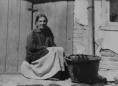 Gelatin silver photograph showing a seated woman beside a woven basket in front of a wooden door and brick wall.