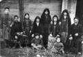 Group of men, women, and children in traditional Tushetian clothing posing outdoors in front of a wooden structure.