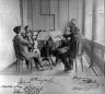 Four men in formal attire seated in a room playing string instruments around music stands.