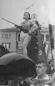 Woman holding a rifle standing on a parade float during a Soviet-era public celebration with crowd and buildings in the background