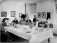 Group of men and women seated around a long dining table inside a large room with framed pictures on the walls and a gramophone visible in the background.