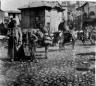 Photograph showing pack animals loaded with goods standing on a cobbled street in the Tiflis bazaar area, with people and carts visible in the background.
