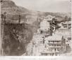 Black and white photograph showing multi-story wooden houses with balconies built on a hillside overlooking a gorge with vegetation.
